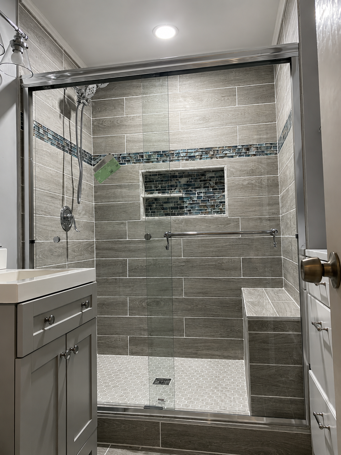 Bathroom with grey wood-look tile shower, glass mosaic accent stripe, built-in niche, and bench seat